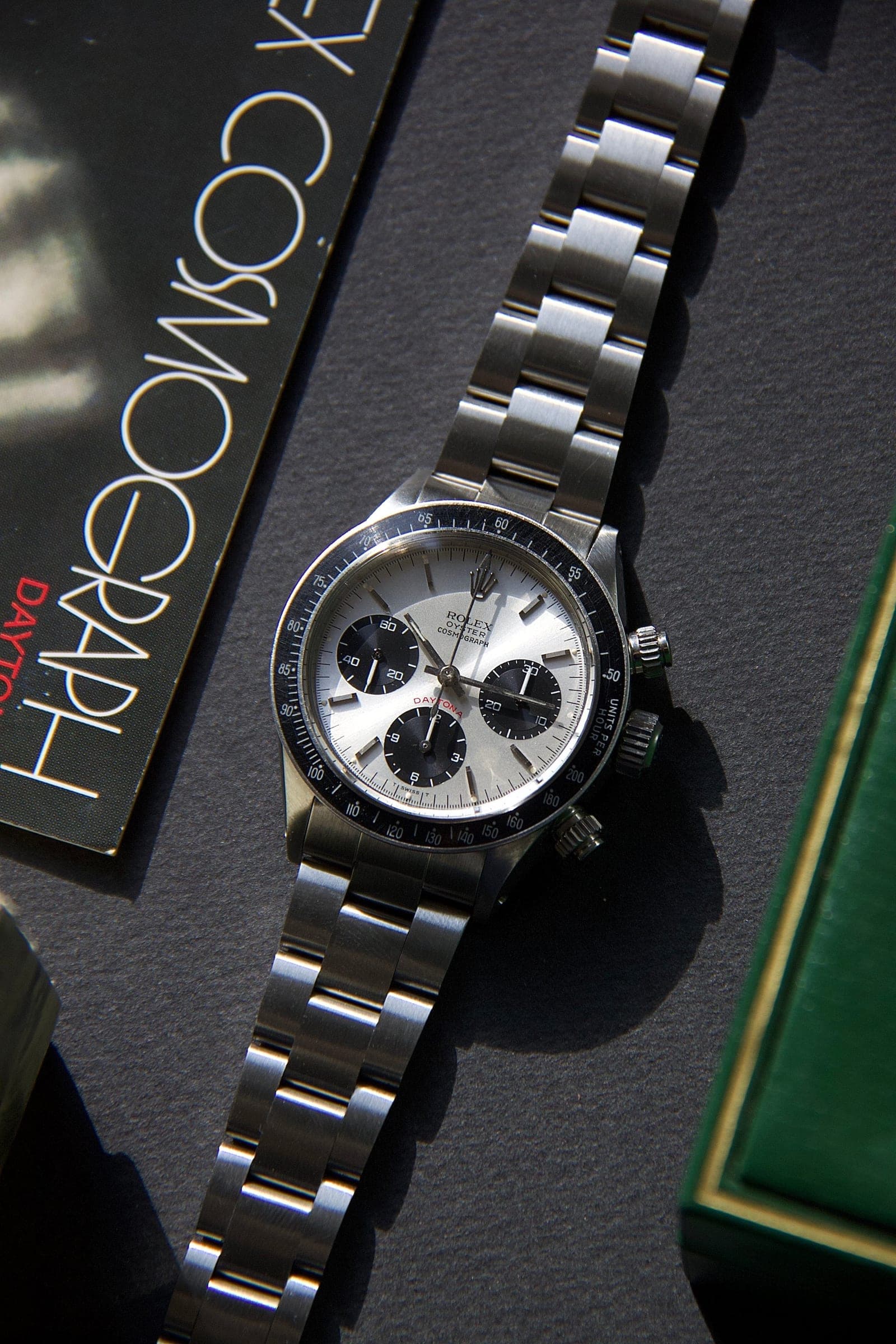 Vintage Rolex Cosmograph Daytona with a silver dial and black sub-dials, beside a Cosmograph booklet and a green presentation box